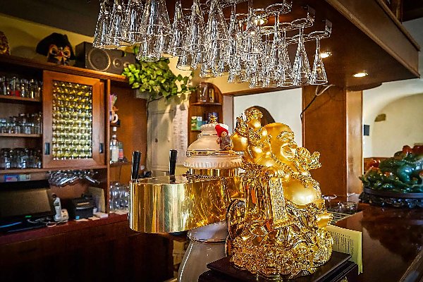 Nahaufnahme einer Bar mit hölzernem Tresen, hängenden Weingläsern und einer goldenen lachenden Buddhafigur im Vordergrund, umgeben von Regalen mit Gläsern und weiteren Dekoobjekten.