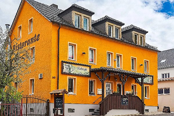 Leuchtend orangefarbenes Gebäude mit der Aufschrift „Ristorante“, dunklem Satteldach, kleinen Dachgauben und einem holzverkleideten Eingang mit Treppe an einer ruhigen Straße.